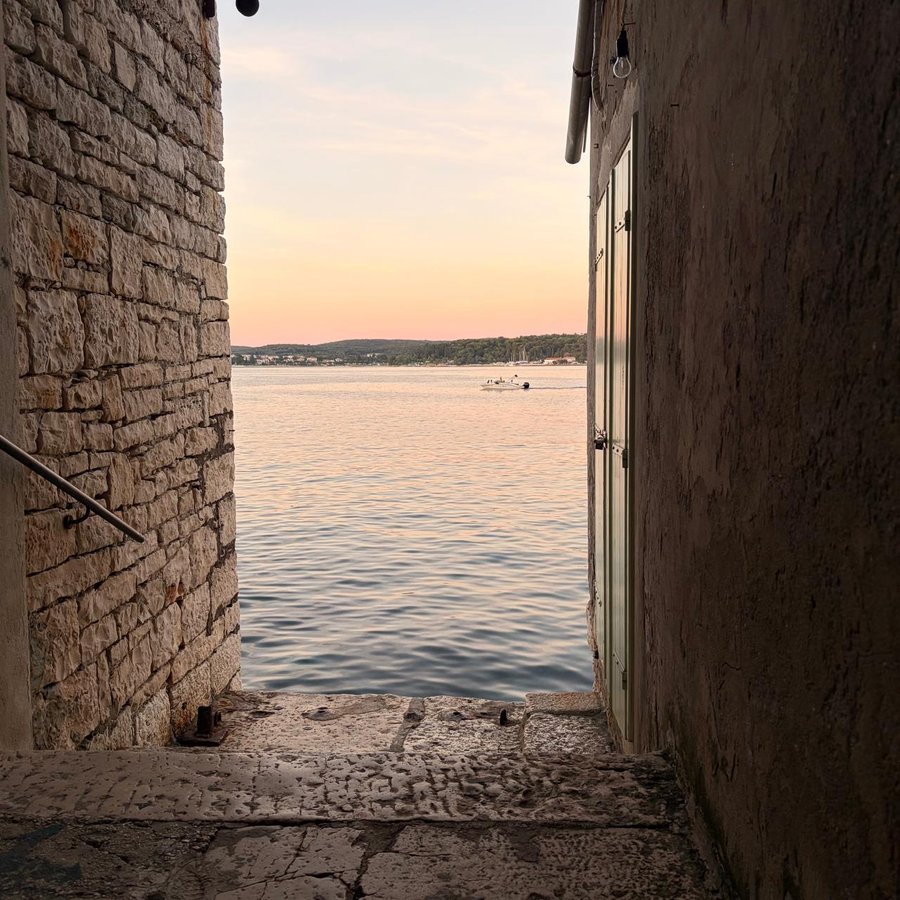 A stone alley opening onto the Adriatic at sunset, with a small boat crossing the water, Rovinj