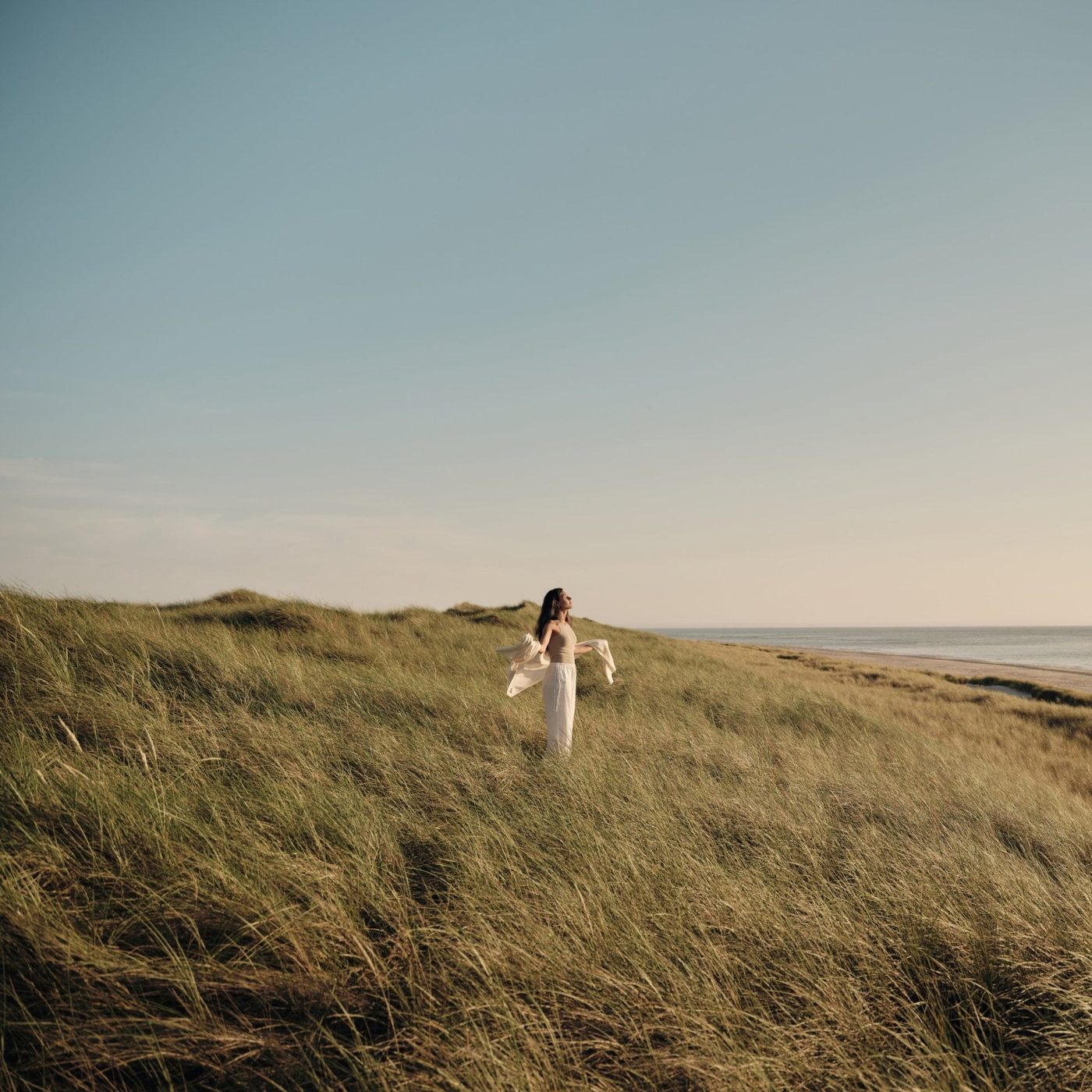 A guest with arms outstretched on a grassy dune above the sea at golden hour, Lanserhof