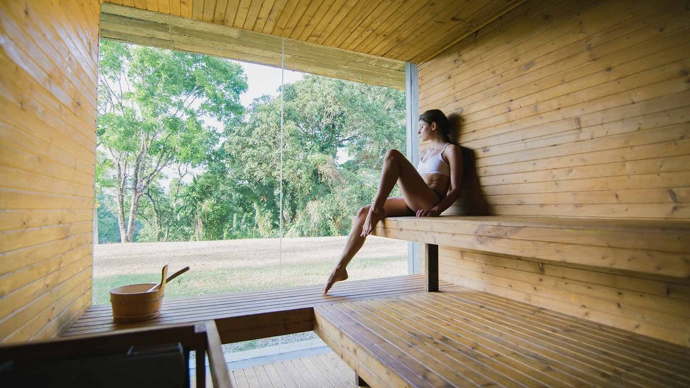 A guest in a glass-walled wooden sauna looking out over the garden, Santani Wellness